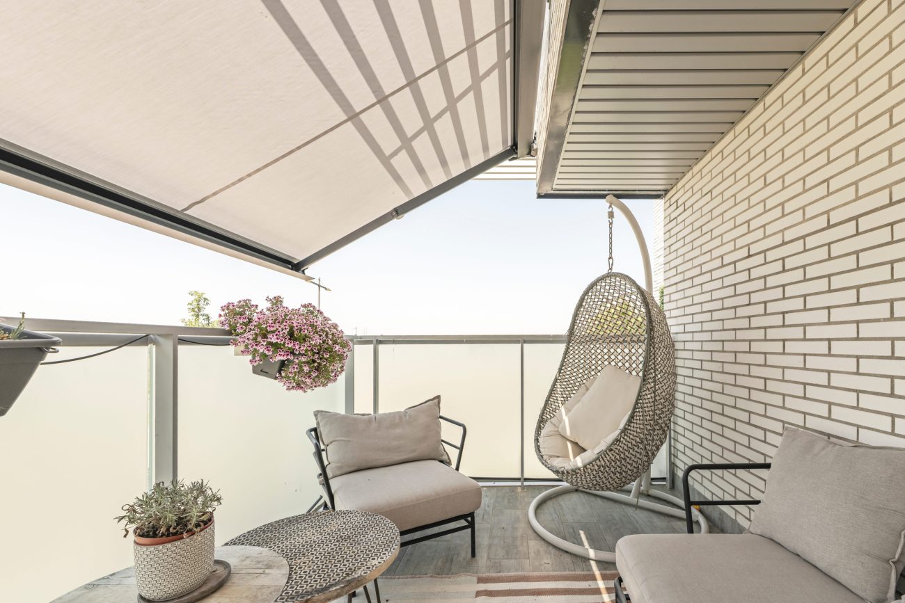 Cozy balcony seating area with a beige cushioned chair, a hanging egg chair, and potted plants by a glass railing.