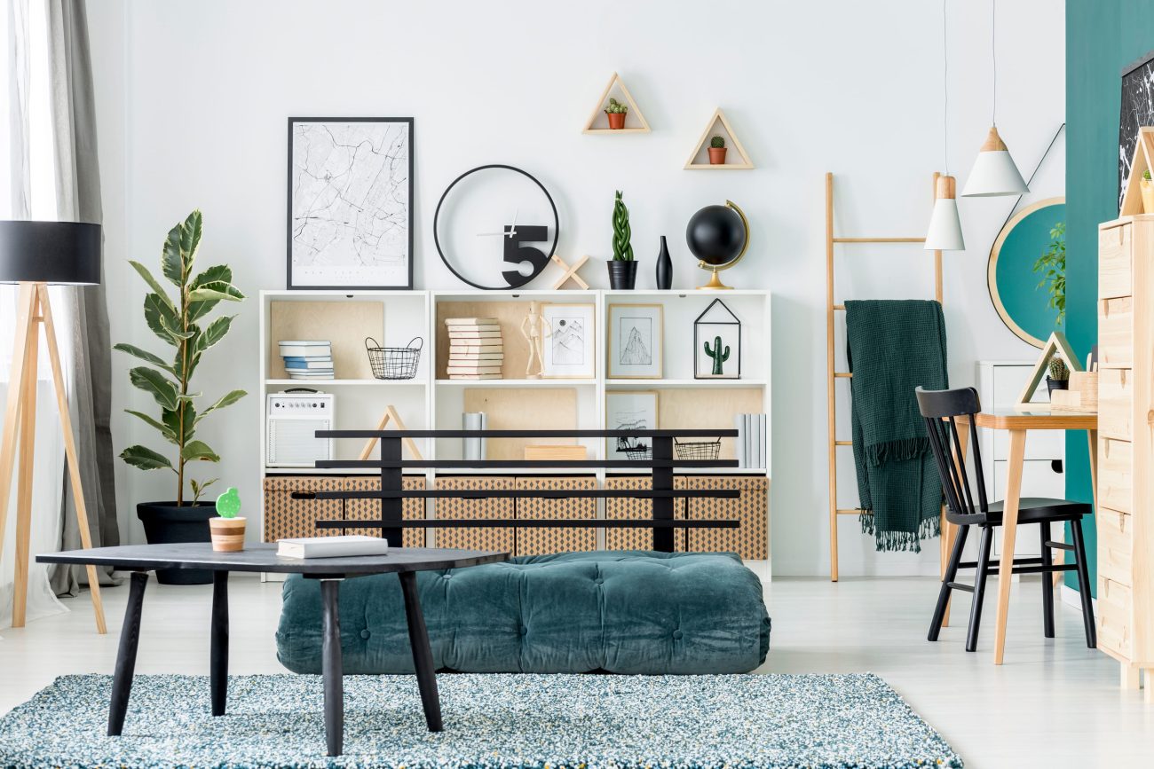 Bright living room with a white shelving unit, teal tufted ottoman, and a black coffee table on a patterned blue rug; plant and decorative items on shelves create a modern, cozy space.