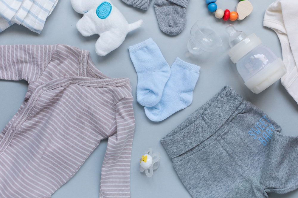 Flat lay of baby items on a blue background: striped onesie, gray pants with 'Sweet Baby' text, blue socks, bottle, pacifier, and a soft toy.
