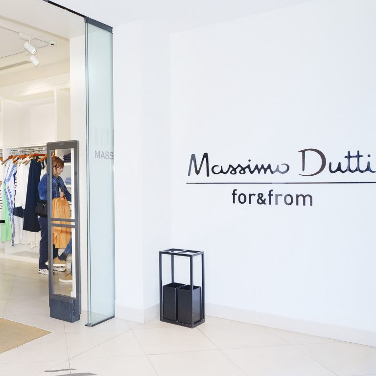 Massimo Dutti store entrance with a glass door, clothing racks visible inside, and a white wall featuring the 'Massimo Dutti for&from' sign.