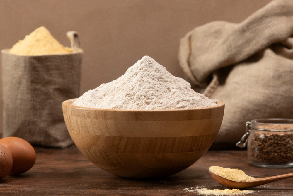 Wooden bowl piled high with white flour on a rustic table, with eggs and a jar of grains in the background.