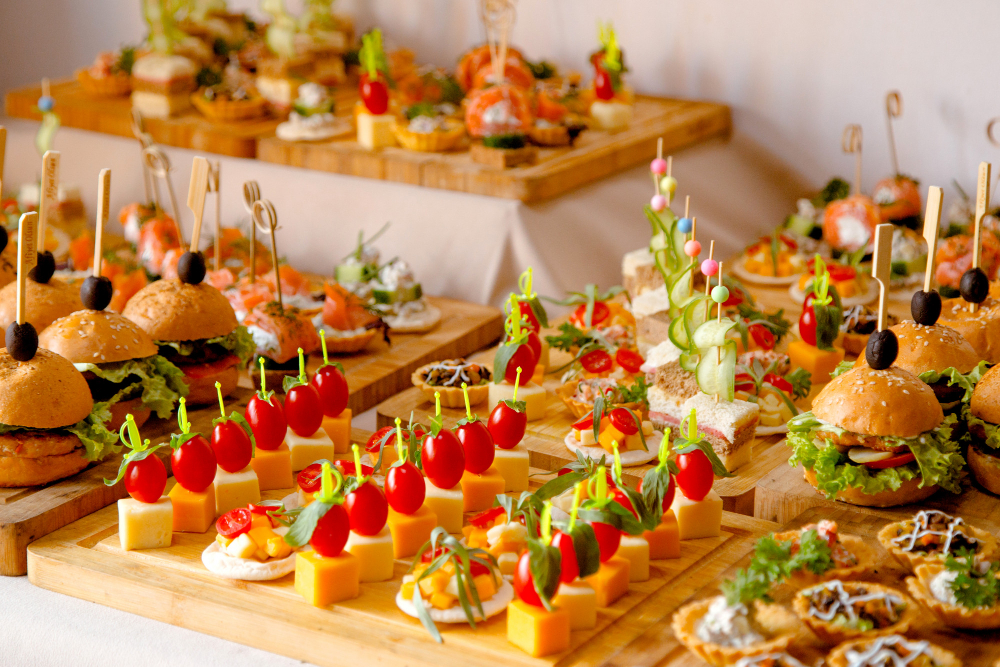 773018 - Pasión por Cocinarte - Diario a pie de barrio Assorted bite-sized hors d'oeuvres on wooden boards, including sliders, cheese cubes with cherry tomatoes, and veggie skewers at a dessert table setup