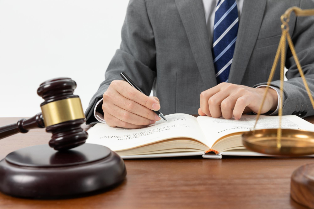 774887 - Rachid M Abogado - Diario a pie de barrio Lawyer in a gray suit signs a book at a desk with a gavel and scales of justice nearby.