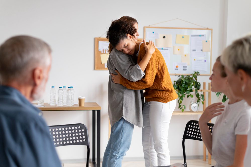 776376 - Mensalud - Diario a pie de barrio Two women share a comforting hug in a bright room during a meeting, while others sit nearby and a bulletin board with notes is in the background.