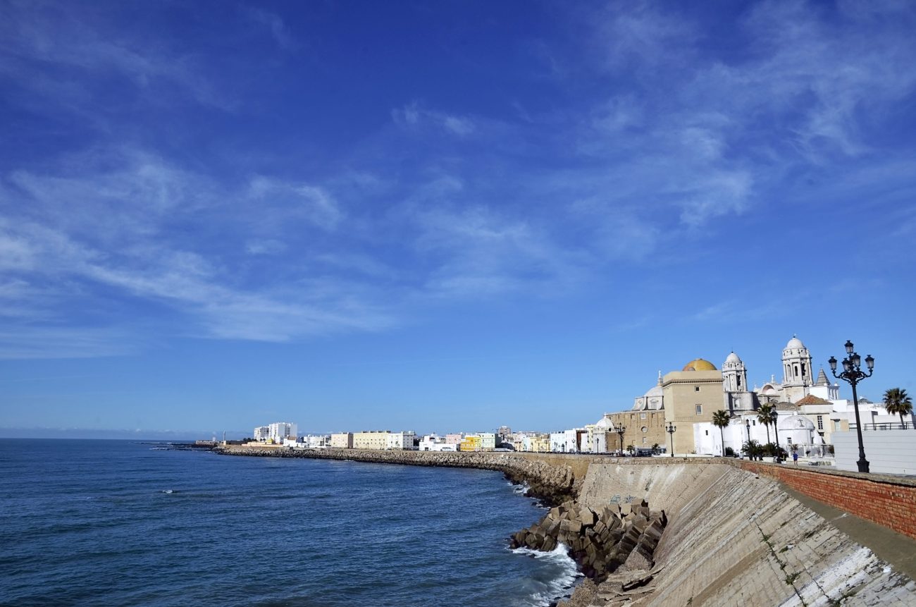 CADIZ SPAIN - Diario a pie de barrio Seaside promenade along a stone seawall with white Mediterranean buildings and a domed church on the right, overlooking the blue sea under a clear sky.