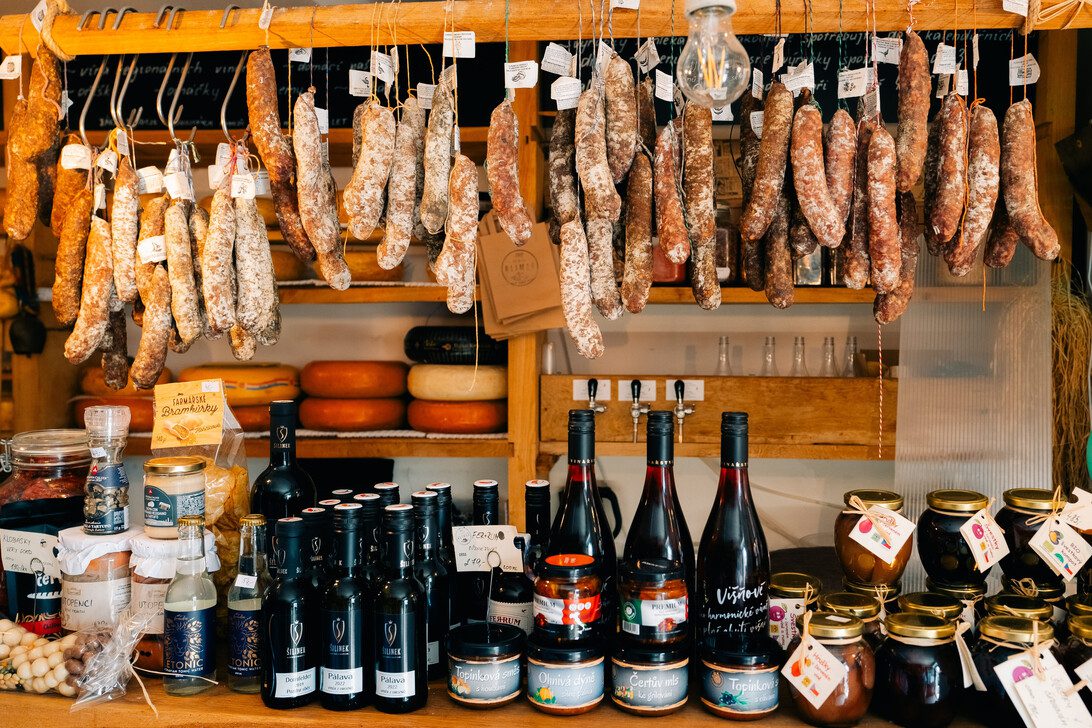 Rustic market stall with rows of hanging cured sausages above a counter of cheeses, bottles, and jars,
