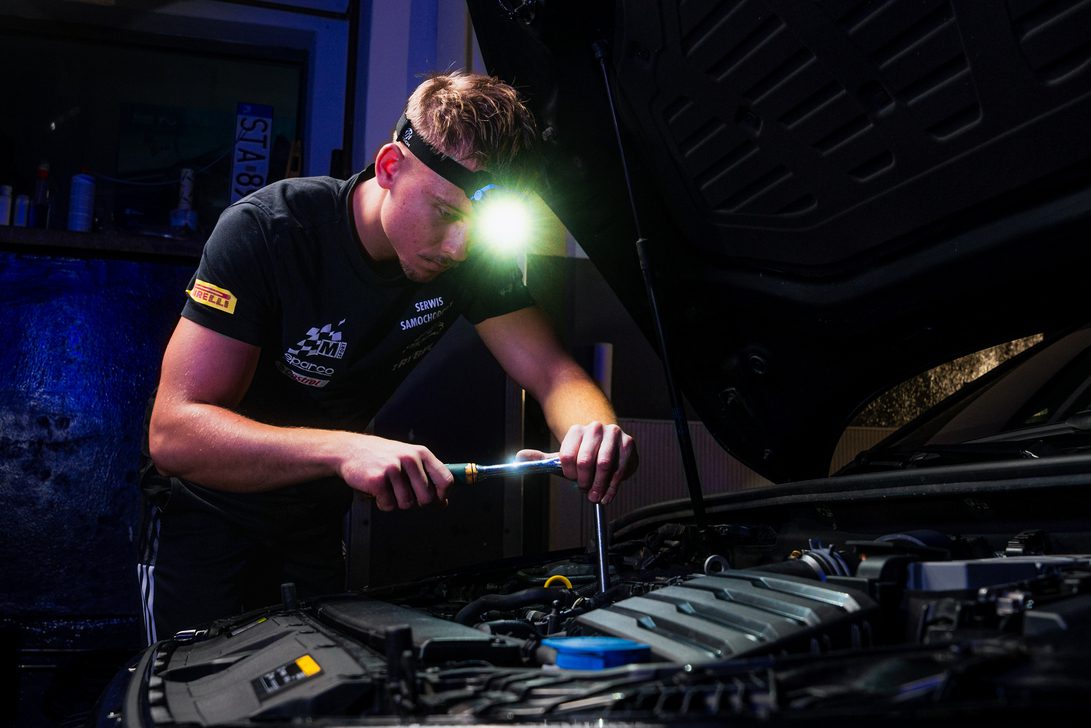 Mechanic wearing a headlamp works under an open car hood, tightening a bolt in the engine bay.