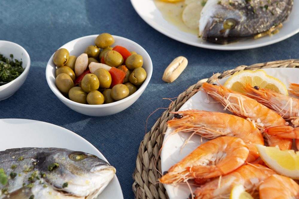 Shrimp platter with lemon wedges served on a white plate and a bowl of olives nearby on a blue table.