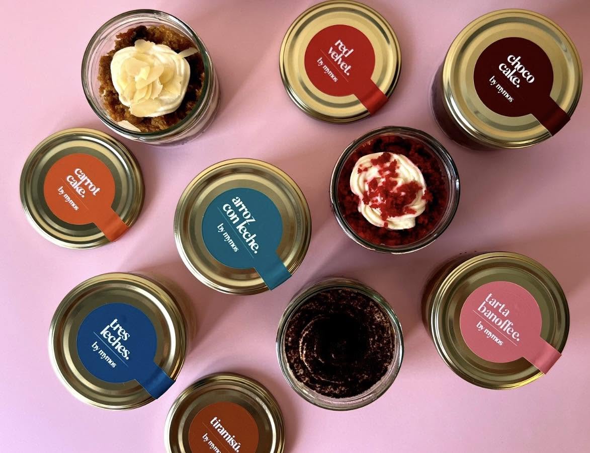 779031 - Mymos - Diario a pie de barrio Top-down view of nine dessert jars with flavored labels on metal lids arranged on a pink background, some with whipped cream toppings.