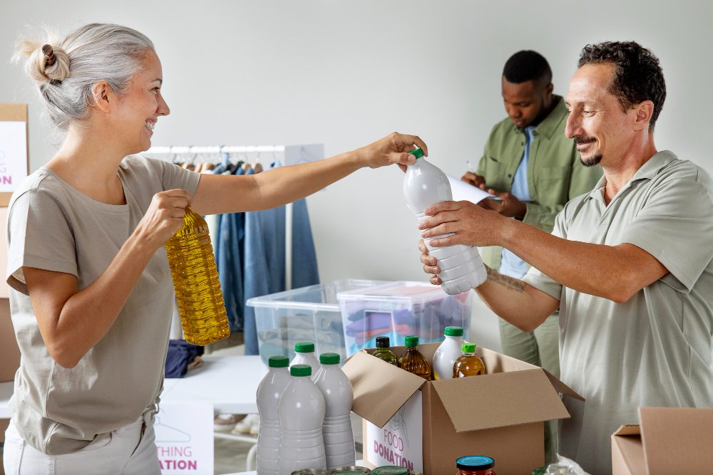780380 - Cáritas Zaragoza - Diario a pie de barrio Two volunteers exchange bottles at a donation event; a woman hands a bottle to a man, smiles all around as boxes and clothing racks appear in the background to donate supplies.