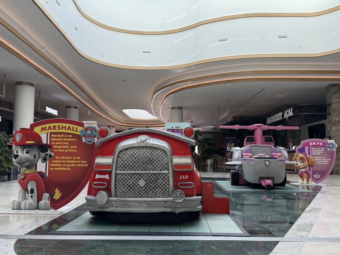 La Patrulla Canina GP2 - IP - Diario a pie de barrio Life-sized PAW Patrol display in a mall: red fire truck centerpiece with Marshall cutout on the left and other toy vehicles nearby.