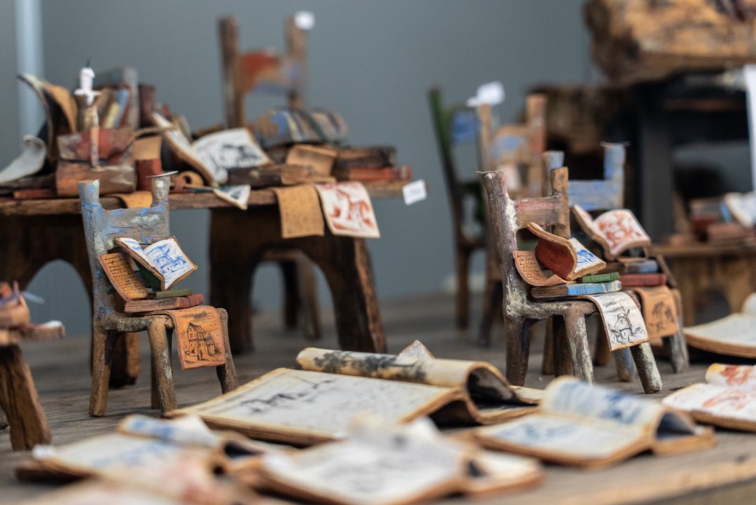 Feria artesania otoño 26 - IP - Diario a pie de barrio Miniature wooden chairs piled with colorful sketches and pages scattered around, as in an art installation.